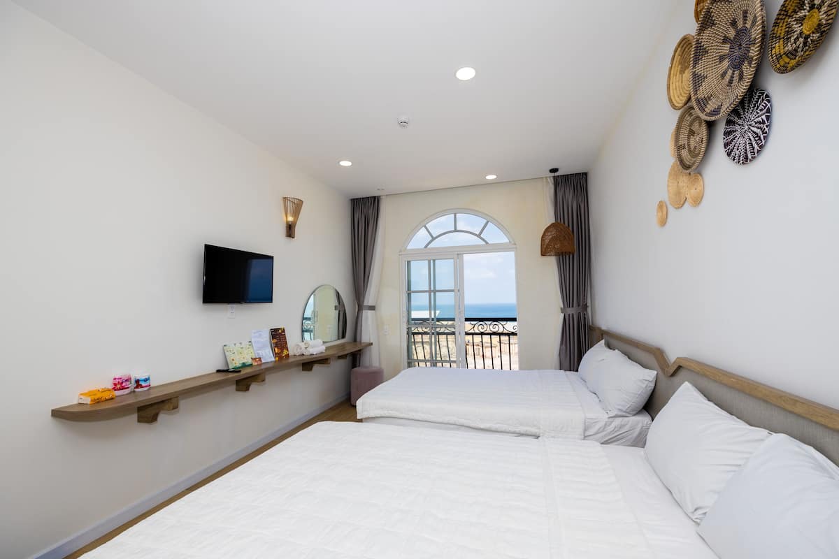 A hotel room with two beds, a TV, a shelf with toiletries, and a balcony with a view of the sea.