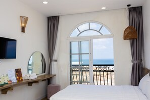 Un cuarto de hotel con cama, televisor, ventana con vista al mar, balcón con barandilla y un televisor montado en la pared.
