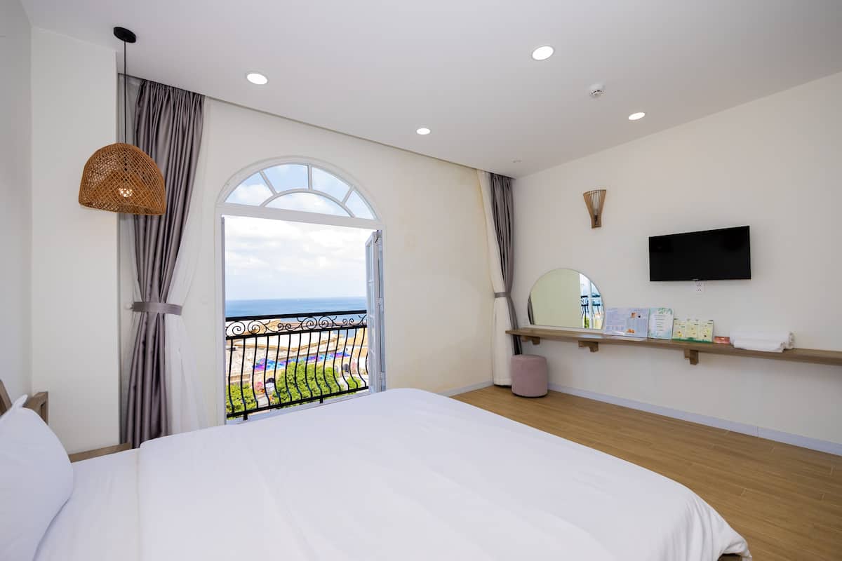 A hotel room with a large bed, a TV mounted on the wall, a small shelf with toiletries, and a balcony with a view of the beach.