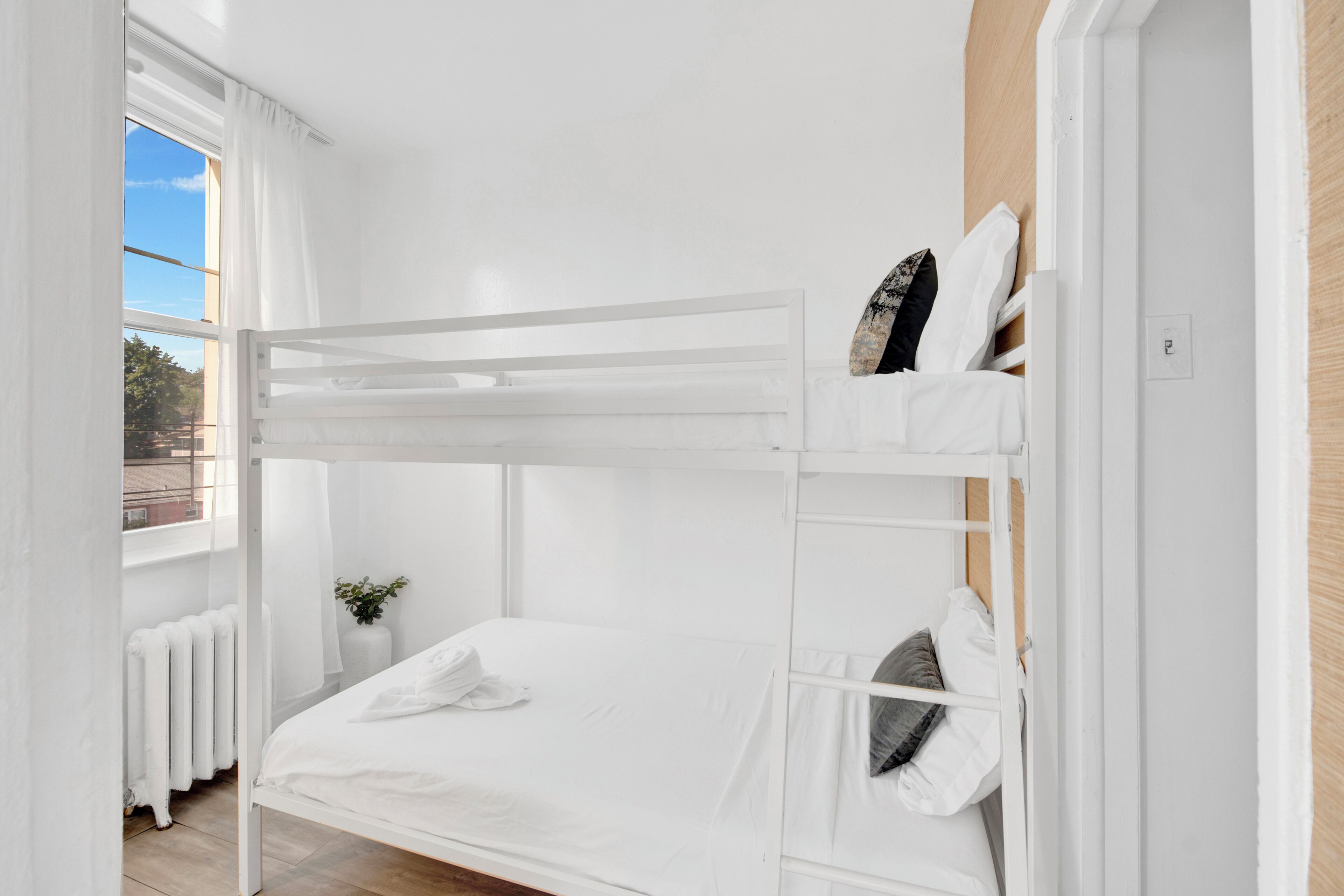 A bunk bed room with white bedding, a small bedside table with a plant, and a window with curtains.