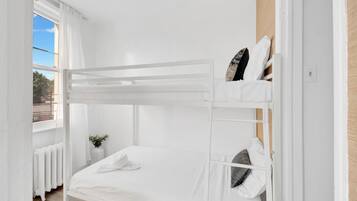A bunk bed room with white bedding, a small bedside table with a plant, and a window with curtains.