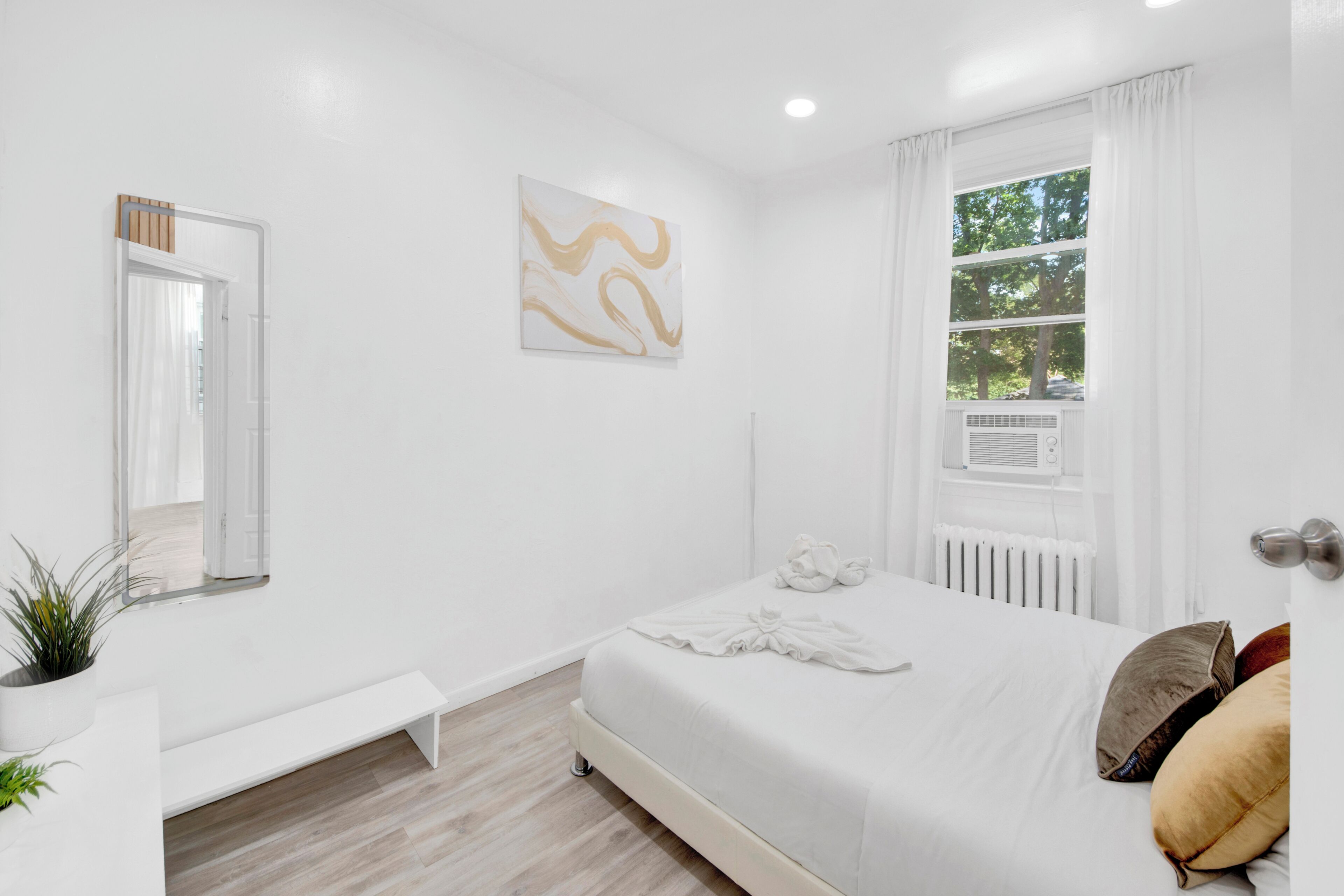 A modern bedroom with a large bed, a bedside table, a bench, a potted plant, and a window with curtains.