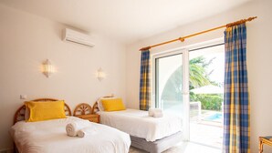 A hotel room with two beds, each with white bedding and a yellow pillow. There are wall-mounted lights, a window with blue and yellow curtains, and a view of a pool outside.