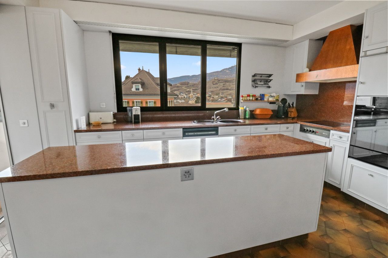 Eine moderne Küche mit einer großen Kochinsel, weißen Schränken und Blick auf ein Haus durch ein großes Fenster.