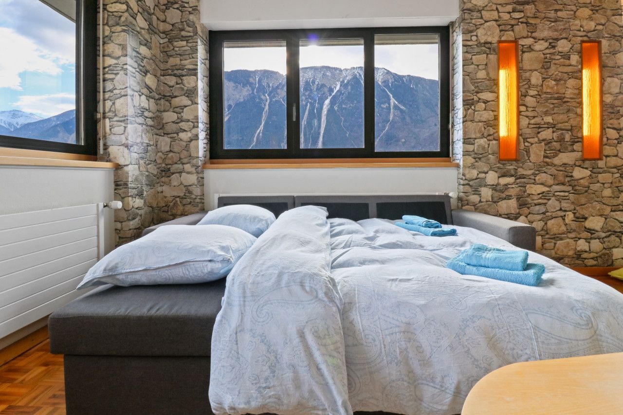 Une chambre avec un mur en pierre, une grande fenêtre donnant sur les montagnes, un lit avec une literie blanche et une table de chevet avec une lampe.