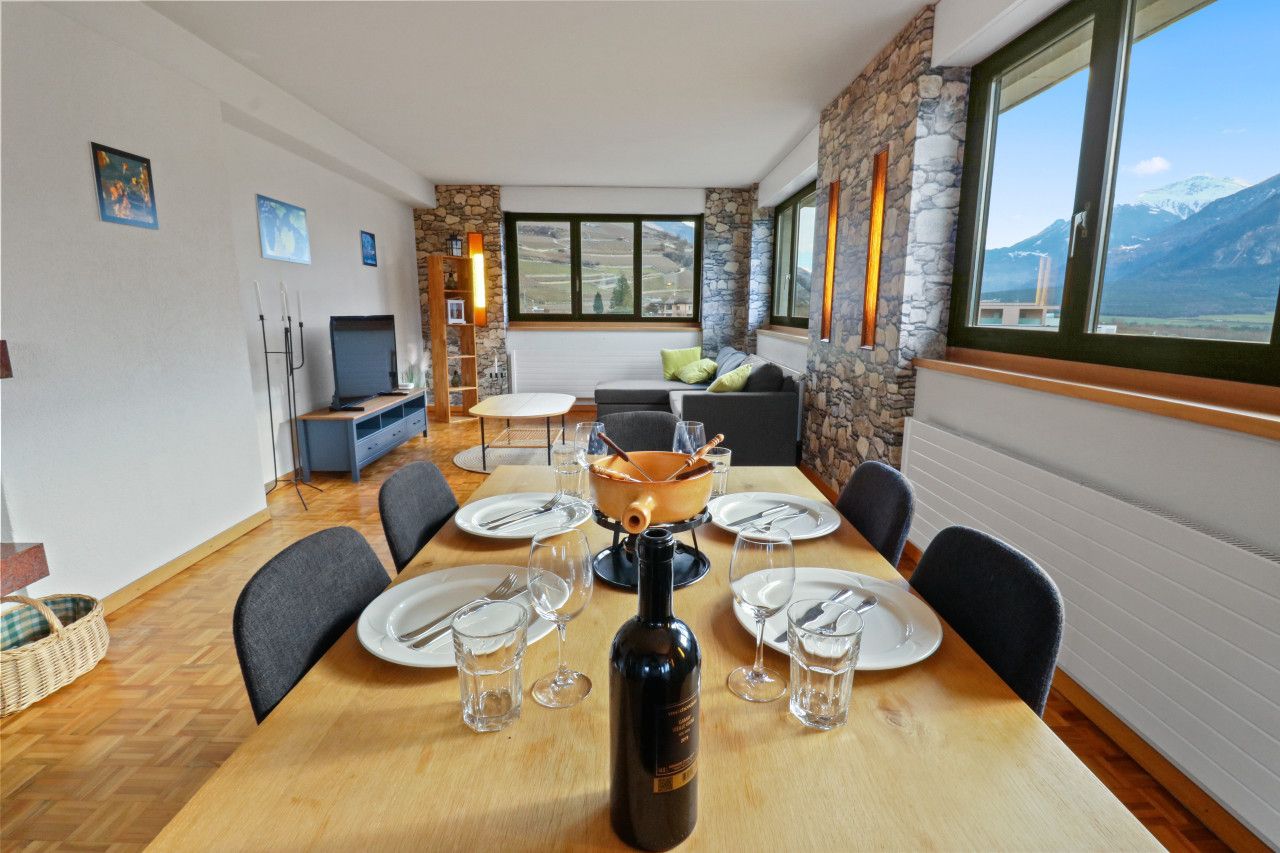 Un coin repas avec une table en bois dressée pour six personnes, une bouteille de vin et une vue sur les montagnes par de grandes fenêtres.