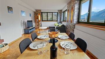 Un coin repas avec une table en bois dressée pour six personnes, une bouteille de vin et une vue sur les montagnes par de grandes fenêtres.