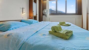 Ein Hotelzimmer mit zwei Betten, einem Fenster mit Blick auf die Berge, einem Standventilator und einer wandmontierten Lampe.
