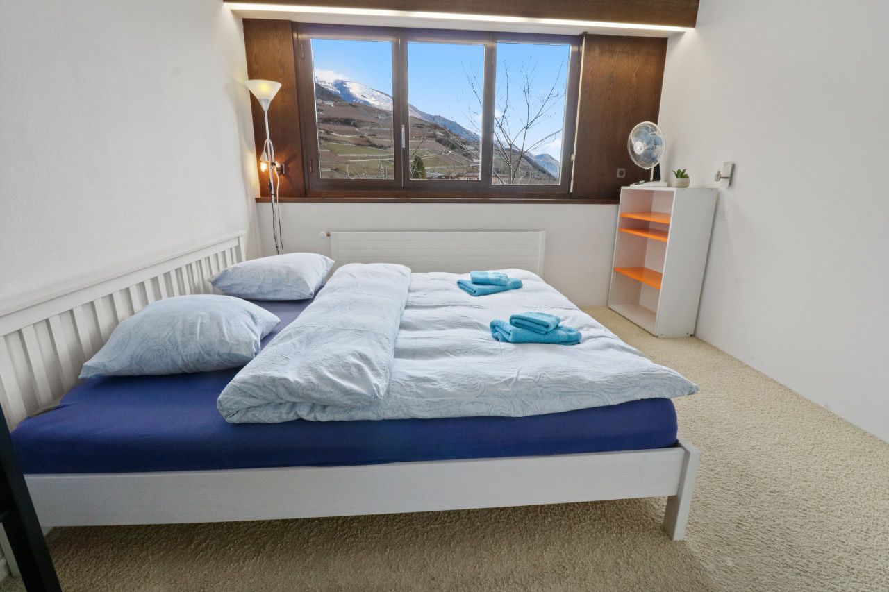 Una camera da letto con un letto, una finestra vista montagna, un comodino e una piccola mensola.