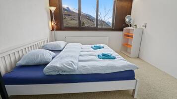 Una camera da letto con un letto, una finestra vista montagna, un comodino e una piccola mensola.
