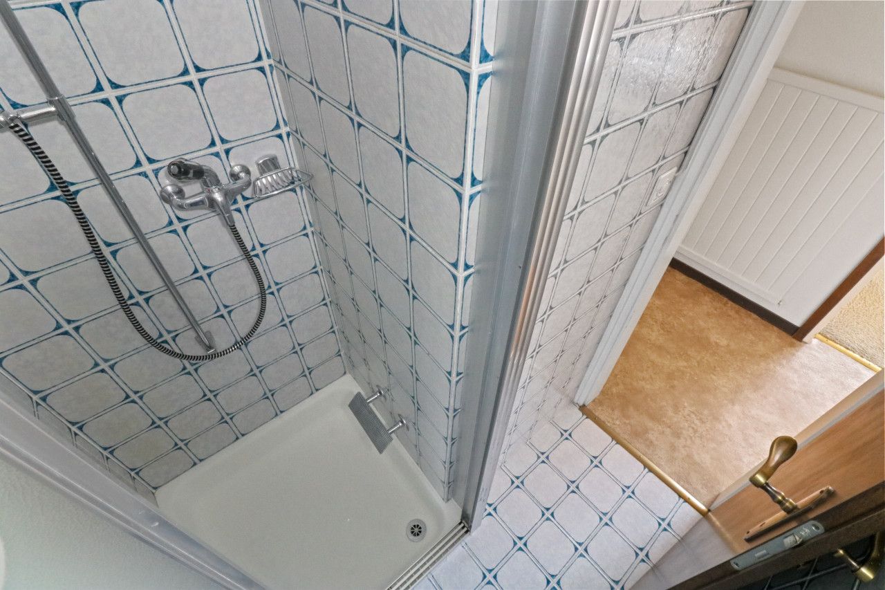 Une douche dotée d’une porte en verre et d’une poignée en métal, des murs carrelés avec un motif géométrique et une porte en bois avec une poignée en laiton.