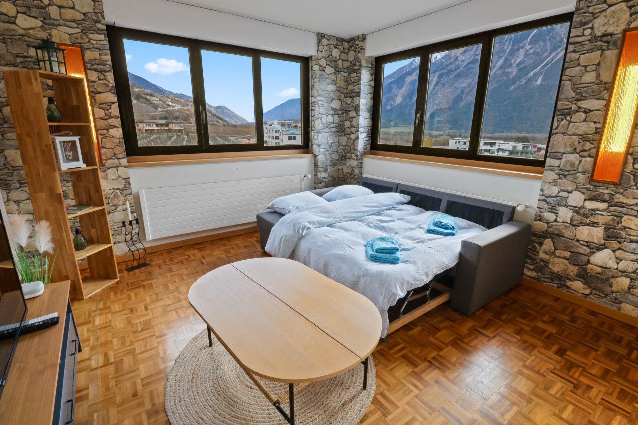 Ein Schlafzimmer mit einem Bett, einem Holztisch, einem Bücherregal und Blick auf Berge durch große Fenster.