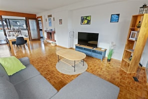 Ein modernes Wohnzimmer mit Holzboden, grauen Sofas, einem Holztisch, einem Fernseher und einem Bücherregal.