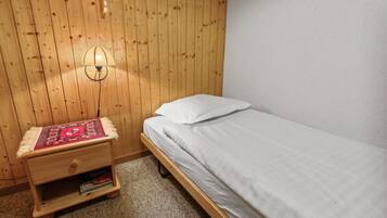 Ein kleines Zimmer mit einem Einzelbett, einem Holzkopfstück, einer Nachttischlampe und einem Nachttisch mit einem rot gemusterten Tuch.