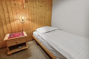 A small, single-bed room with a wooden headboard, a bedside lamp, and a nightstand with a red-patterned cloth.