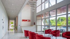 Un hall d’hôtel moderne avec un long couloir, des fauteuils rouges, un comptoir d’accueil et de grandes fenêtres donnant sur les voitures garées et la verdure à l’extérieur.