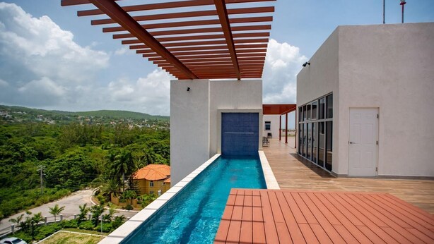 Une piscine extérieure moderne, dotée d’une terrasse en bois, d’un espace abrité avec des poutres rouges et offrant une vue sur un paysage verdoyant luxuriant.