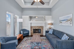 Un salon doté d’une cheminée, d’un ventilateur de plafond et d’une grande fenêtre avec des stores.