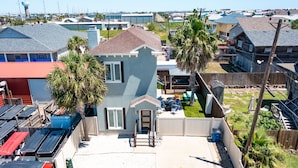 A residential area with a house featuring a front porch, a garage, and a fenced yard with palm trees. - Retreat Port Aransas | A Retreat DFW Experience (Port Aransas)