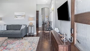 A modern living room with a gray sofa, a coffee table, and a patterned rug. There is a wooden bench with a glass container and a small table with a lamp. A TV is mounted on the wall, and a door with a glass panel is visible. - Retreat Port Aransas | A Retreat DFW Experience (Port Aransas)