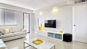 A modern living room with a sofa, a coffee table, a TV mounted on the wall, and a white door. - golden isles # 509 Water view (Hallandale Beach)