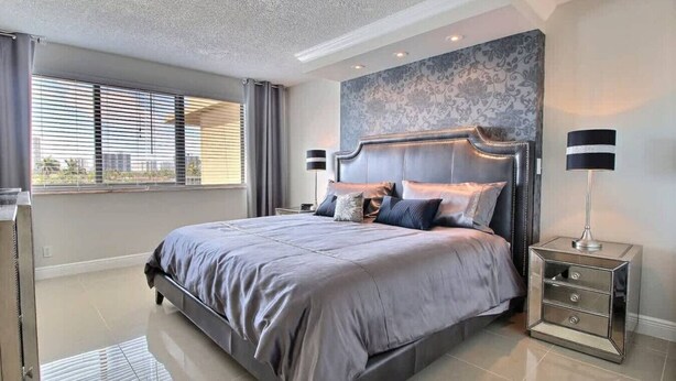 A modern bedroom with a large bed, a headboard with built-in lighting, a nightstand with a lamp, and a view of the outside through the window. - golden isles # 509 Water view (Hallandale Beach)