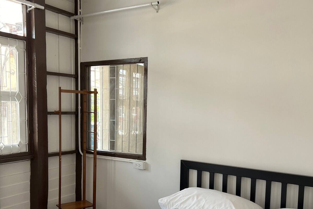 A neatly made bed with white linens and two rolled towels, a wooden ladder leaning against a wall, and a window with bars.