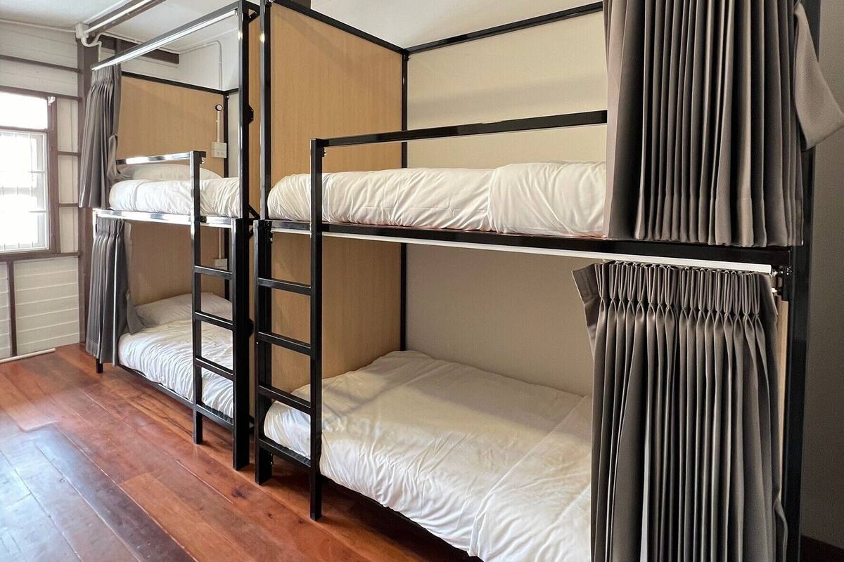 A bunk bed room with wooden flooring, a window with curtains, and a white wall.