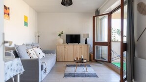 A modern living room with a grey sofa, a wooden cabinet, a TV, and a large sliding glass door to the outside. - Studio Les Vacances a Andernos (Andernos-les-Bains)