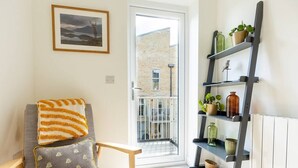 Una habitación con una silla, un almohadón, una estantería con plantas, un cuadro enmarcado y vistas a través de una puerta.