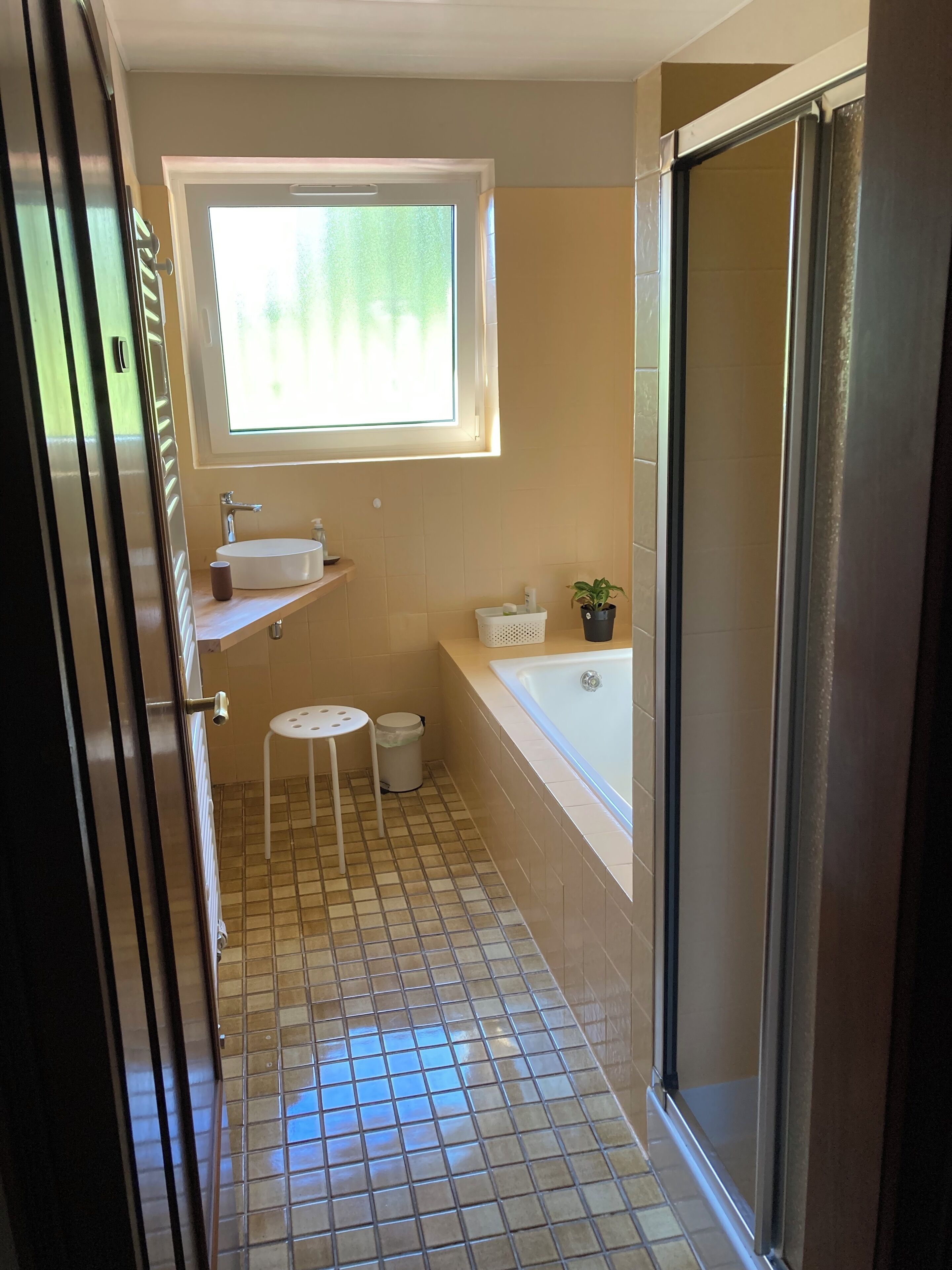 Une salle de bain avec une fenêtre, un lavabo, des toilettes, un petit tabouret et un espace douche.