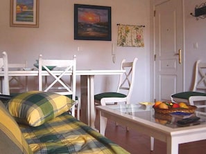 A room with a green and yellow plaid sofa, a glass-top dining table with chairs, a painting of a sunset, and a door with a brass handle.