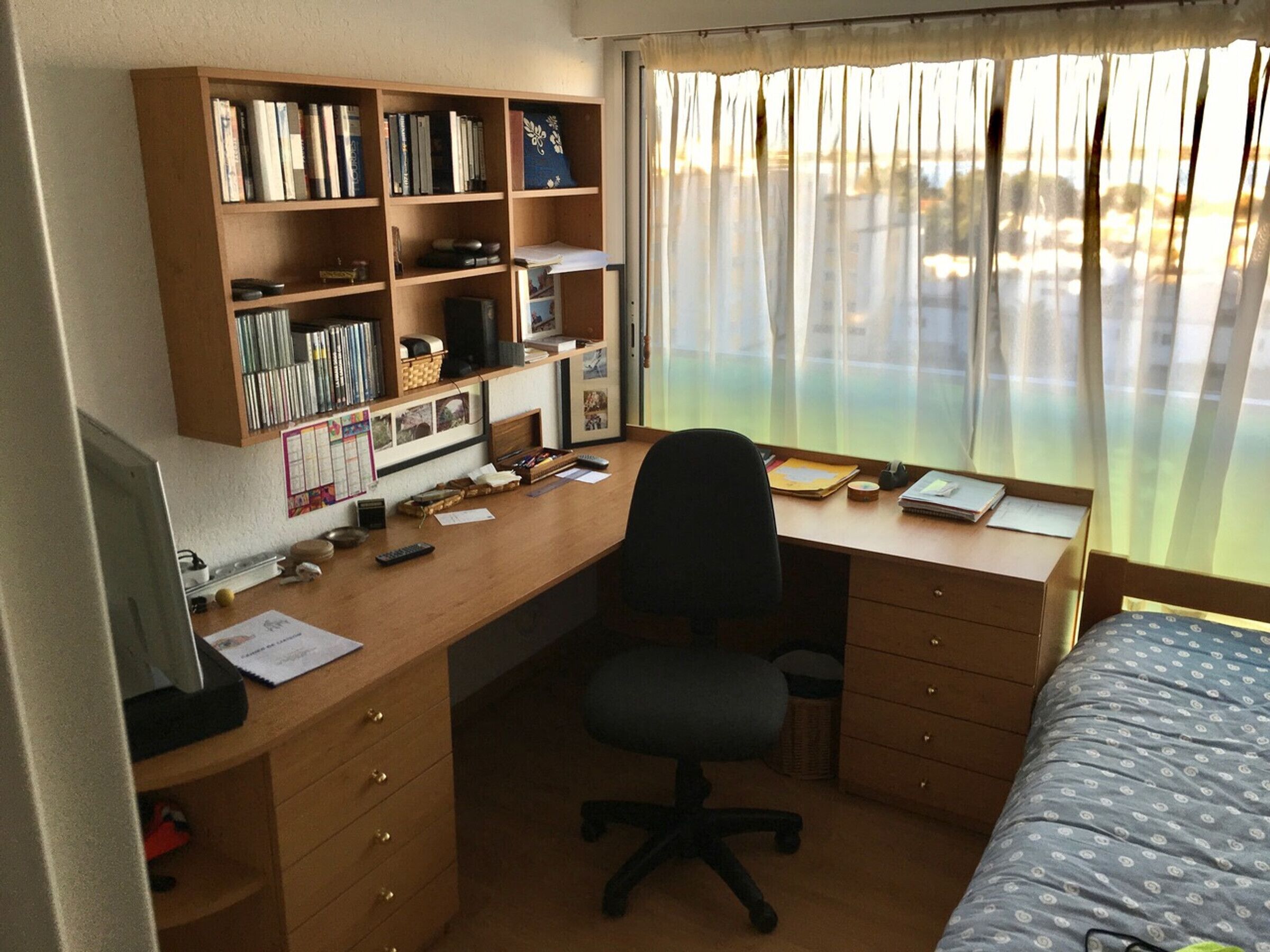 Una habitación con escritorio de madera, computadora, silla, cama con ropa de cama estampada, estantería y ventana con cortinas.