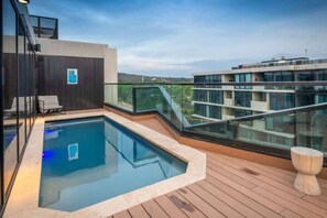 Una piscina en la azotea con barandilla de cristal, terraza de madera y vistas de edificios circundantes.