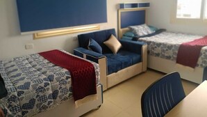 A room with three beds, each with a different headboard design. There is a desk and a chair in the center of the room. - MADEII project training center hostel (Al-'Amriyah)