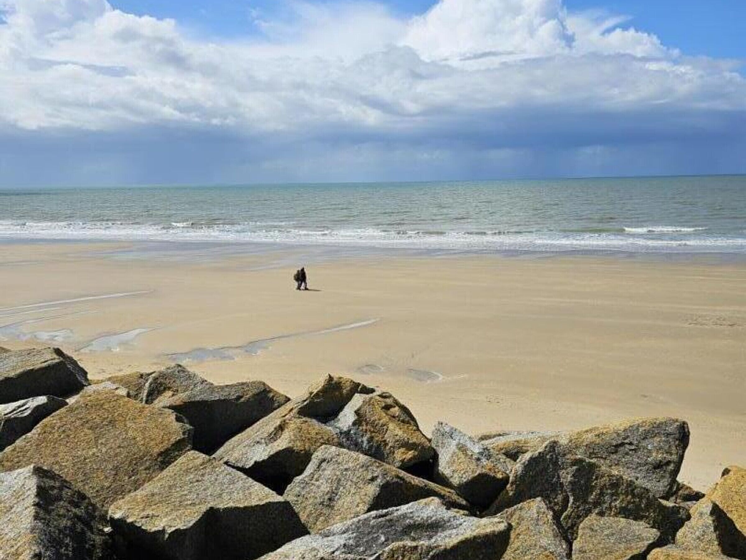 A beach with large rocks in the foreground and a person walking on the sand.
