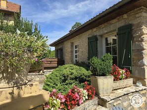 A stone building with green shutters, surrounded by lush greenery and flower beds.