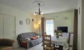 Un salon avec un canapé, une table à manger et des chaises, un lustre et un téléviseur en arrière-plan.