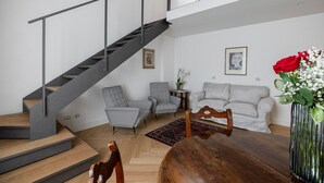 A modern living room with a staircase, grey armchairs, a white sofa, and a wooden dining table.
