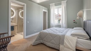 A bedroom with a bed, a bedside table, a nightstand, a window with curtains, and a bathroom with a shower and sink.