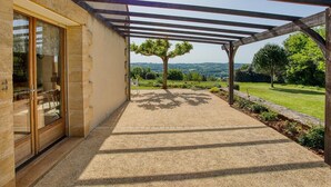 Un patio couvert agrémenté d’une pergola, une entrée avec une porte vitrée et une vue sur un arbre et le paysage environnant.