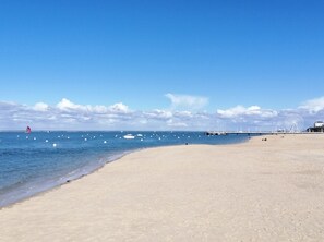 A beach with a clear sky, calm sea, and a distant pier with a large white wheel structure.