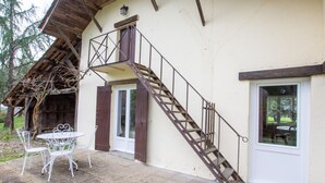 A rustic building with a wooden staircase leading to a door, a small outdoor seating area with metal chairs, and a window overlooking a garden.