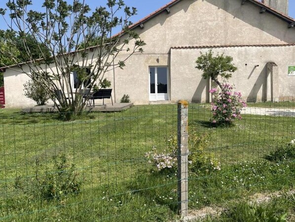 A building with a door, a bench, and a fenced yard with grass and a tree.
