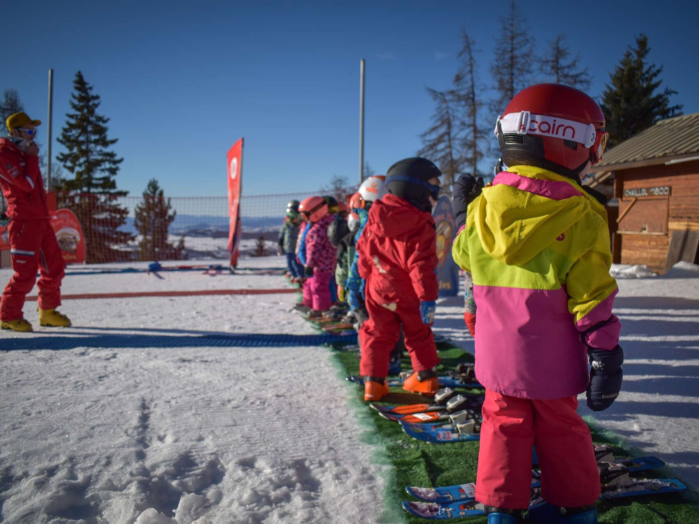 A group of children in colorful winter clothing lined up on a snowy surface, possibly preparing for a skiing activity.