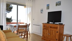 A living room with a sofa, a TV on a wooden cabinet, a dining table with chairs, and a view through a large window.