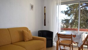 A room with a mustard-colored sofa, a black chair, a wooden dining table with a white tablecloth, and a window with a view of trees.