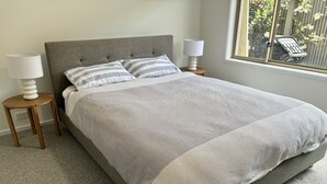 Una cama bien tendida con un cabecero gris, dos mesitas de noche con lámparas, y una ventana con vistas a la vegetación.