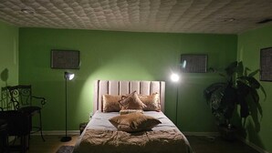 A bedroom with a green wall, a tufted headboard, a bed with multiple pillows, two wall-mounted air vents, a floor lamp, a desk with a chair, and a potted plant.