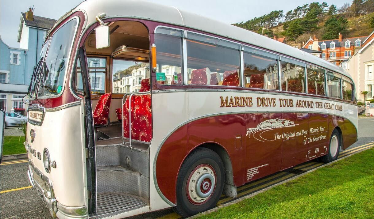 En ældre bus med teksten "MARINE DRIVE TOUR AROUND THE GREAT ORME" på siden, parkeret på en gade med bygninger i baggrunden.